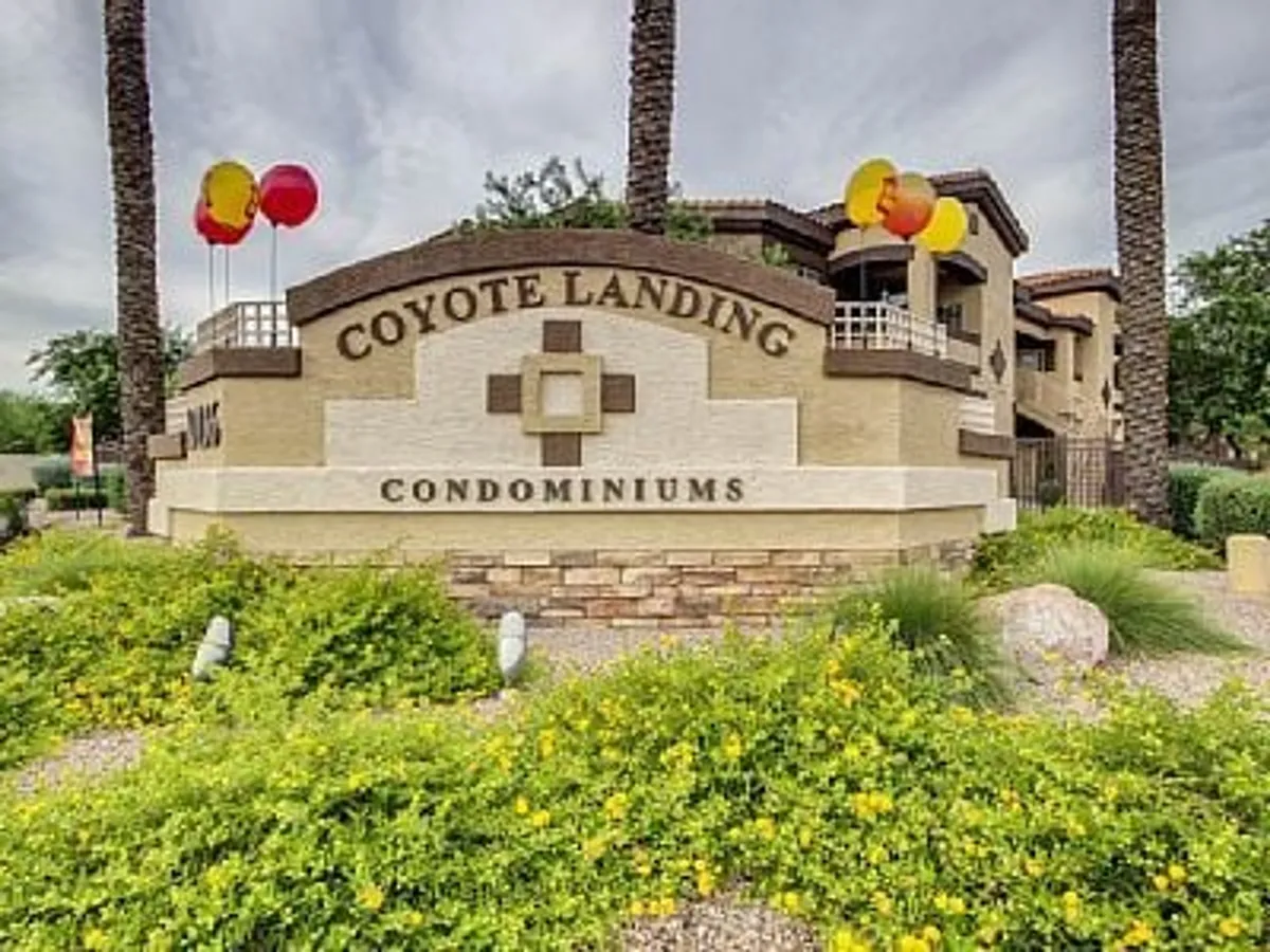 The entrance of Coyote Landing is adorned with festive balloons, a prominent community sign, palm trees, and vibrant landscaping. The welcoming scene radiates community spirit and the joy of arrival.