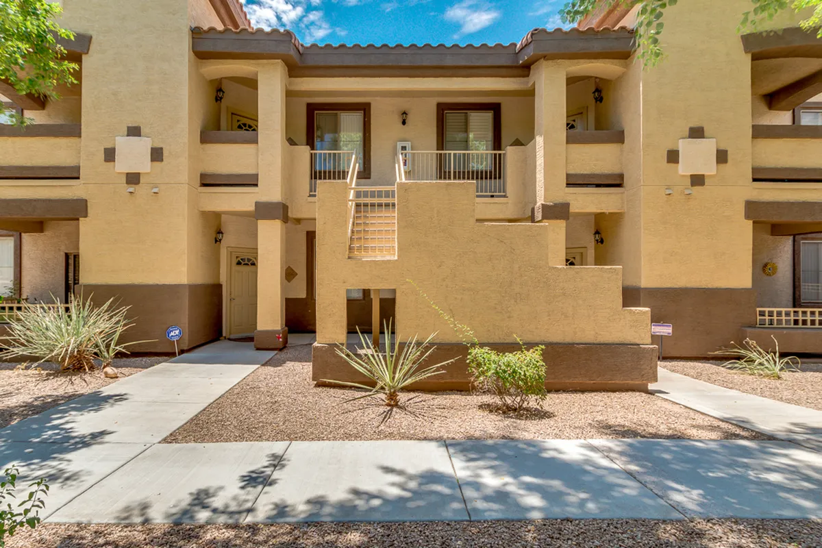 A staircase leading to the upper floor of the condo complex, surrounded by desert landscaping and architectural features. The scene is peaceful, showing the building's exterior and the accessibility via stairs.