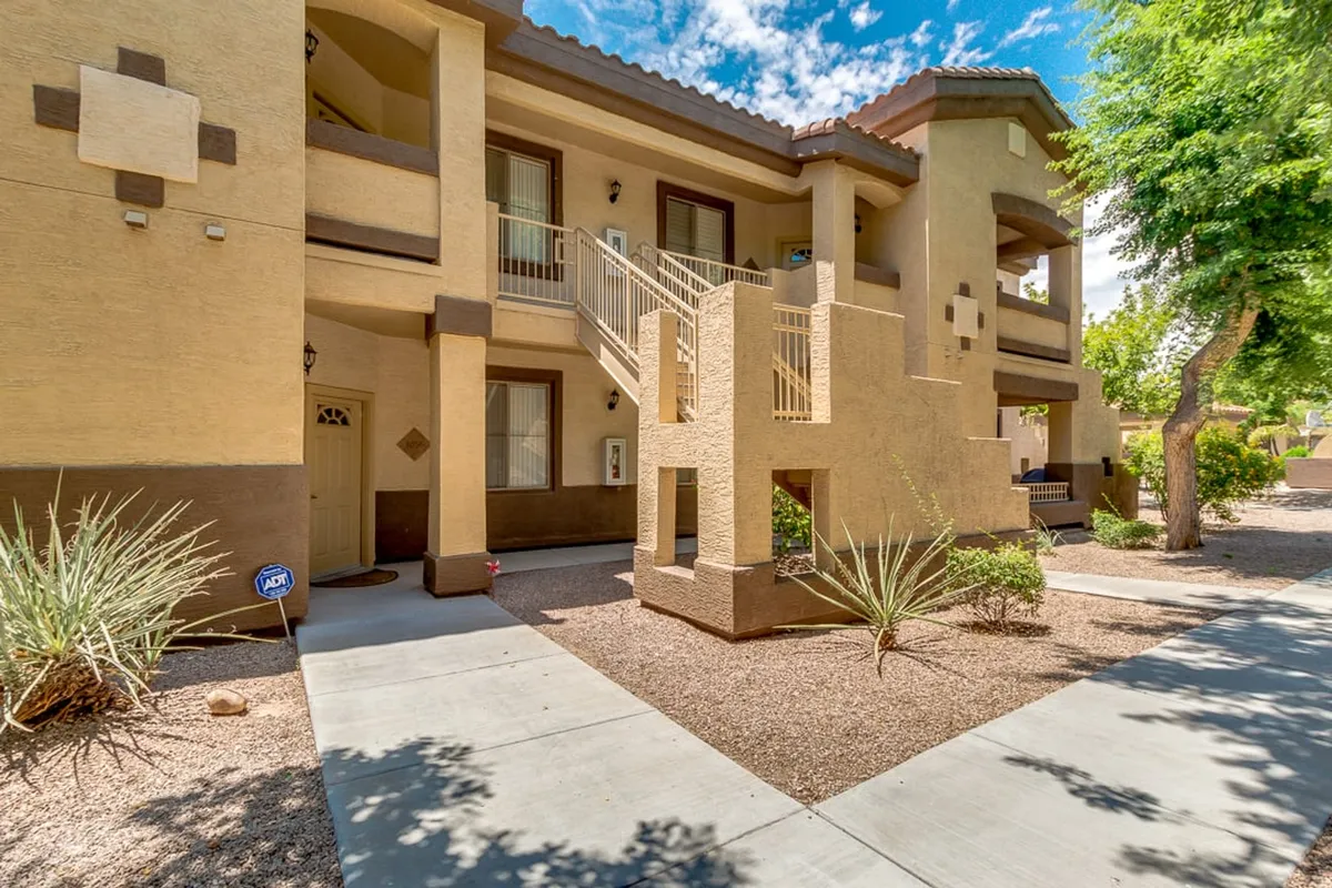 A welcoming exterior view features a staircase, entrance door, manicured shrubs, and the building’s exterior walls. The landscaped surroundings and communal entrance evoke a sense of arrival, safety, and community connection.