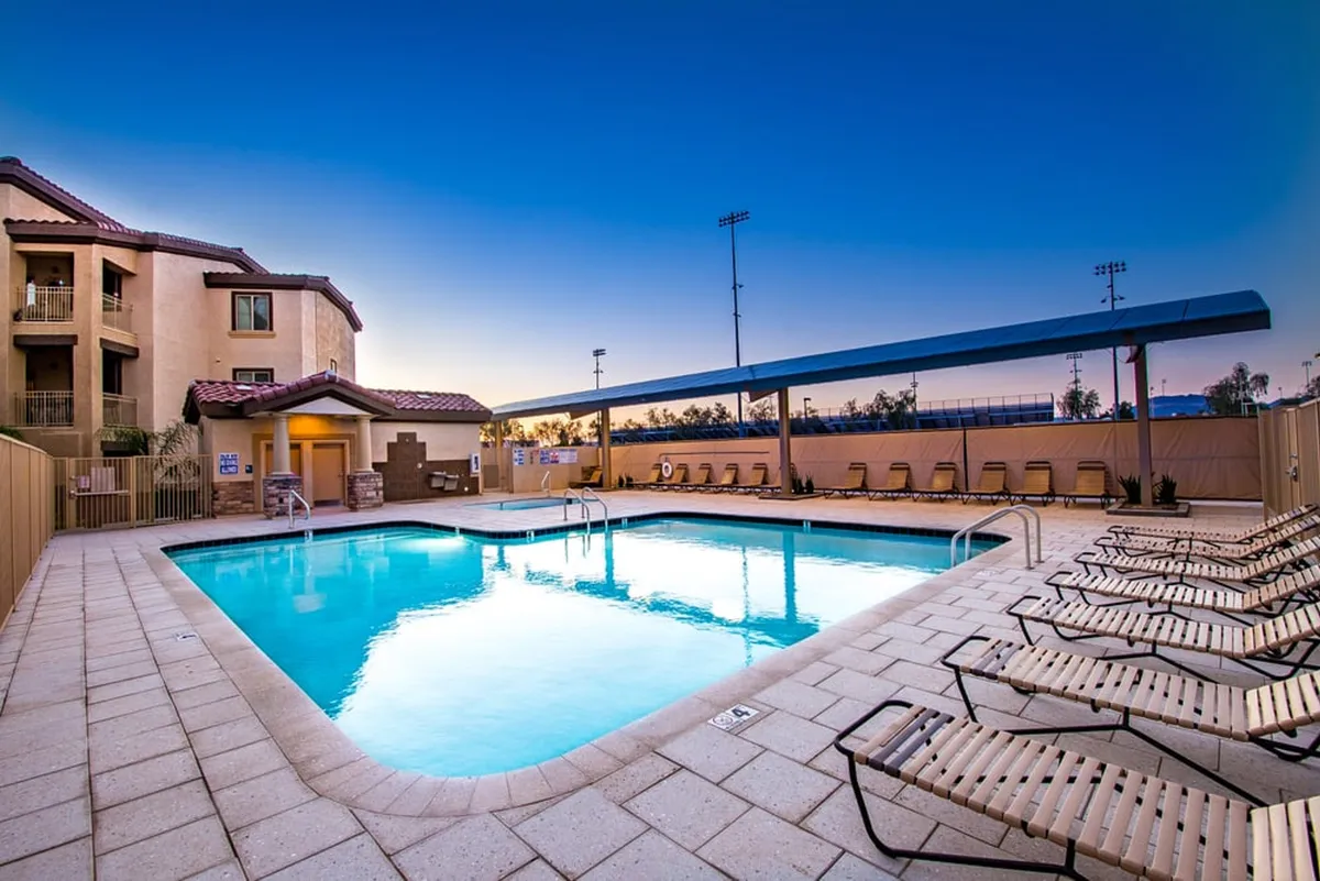 This image captures a sunlit swimming pool area with inviting lounge chairs, a shade structure, and the building's facade in view. The pool and seating are arranged to encourage relaxation and social gatherings, set against a backdrop that hints at a restful retreat.
