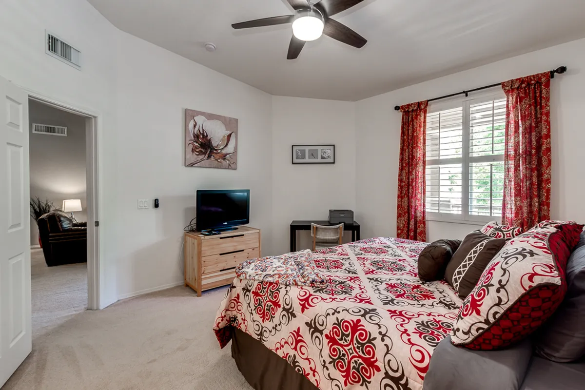A guest bedroom with a patterned queen bed, a TV stand with television, and a window letting in natural light through curtains. The setup is inviting and perfect for leisure or watching TV from bed.