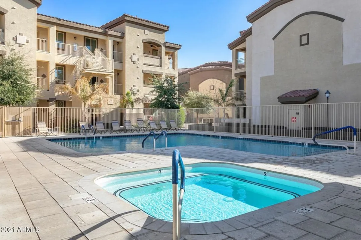 This exterior image captures the inviting community pool surrounded by lounge chairs, a jacuzzi, lush greenery, and the resort-style architecture of the buildings. The tranquil water, ample seating, and palm trees evoke a relaxing, vacation-inspired atmosphere.