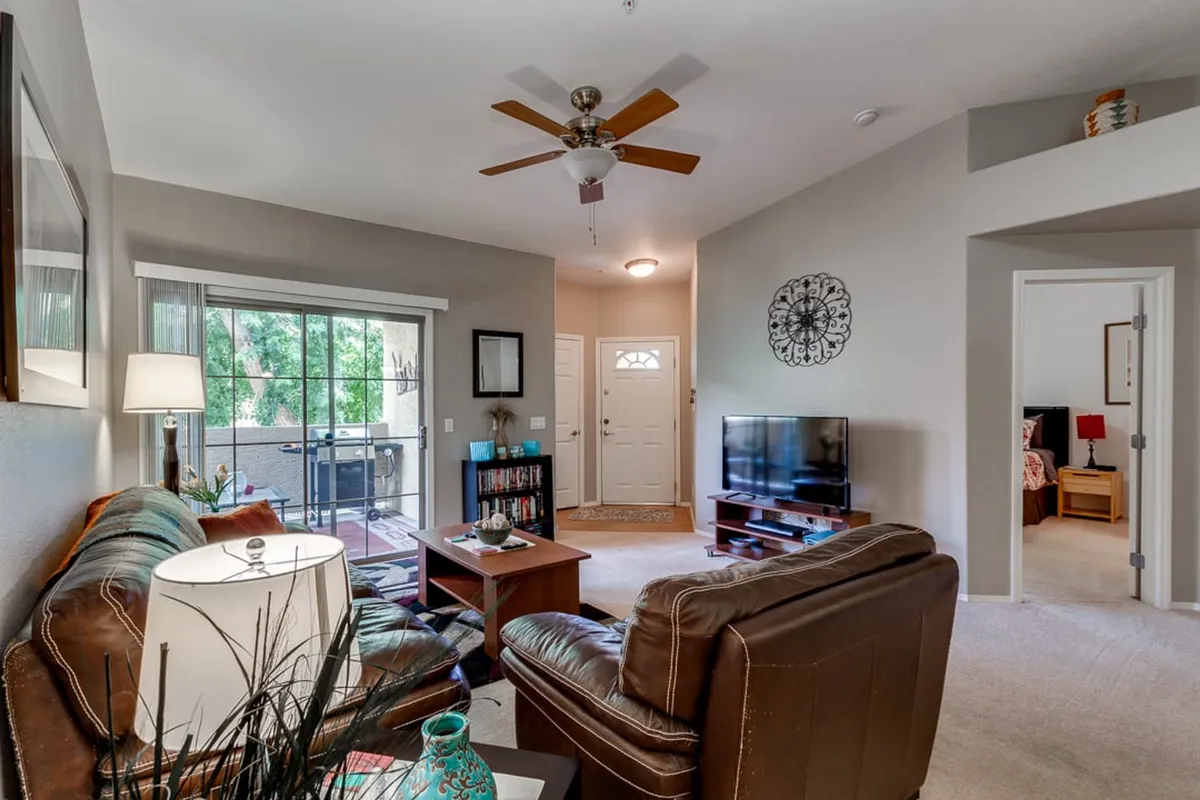 A cozy living room featuring a leather sofa, coffee table, and a large television. Natural light floods the space through a glass door that leads to the patio, offering views of lush greenery outside. The décor is tranquil and modern, with neutral tones and inviting textures.